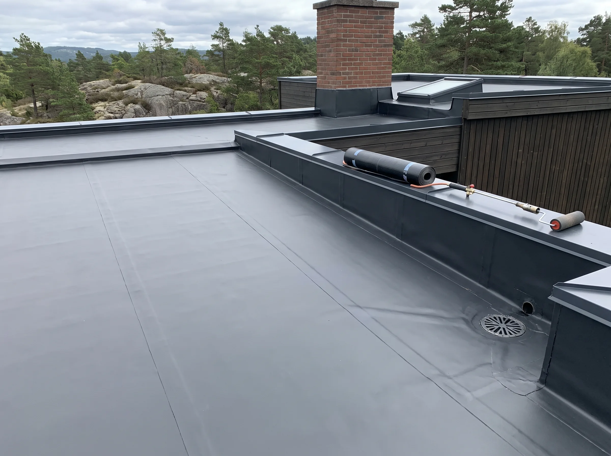 Flatt tak med takpapp og pipe, omgivelse med trær i bakgrunnen